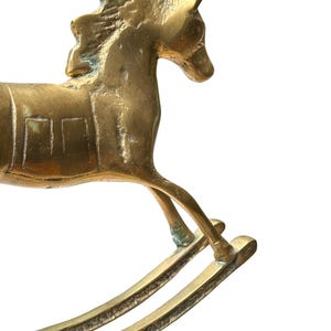 Puede incluir: Una figura de caballo balanc&iacute;n de lat&oacute;n con un acabado dorado met&aacute;lico que presenta p&aacute;tina y oxidaci&oacute;n visibles. El caballo est&aacute; representado en una pose de galope con crines texturizadas, un detalle de silla de montar y dos balancines.
