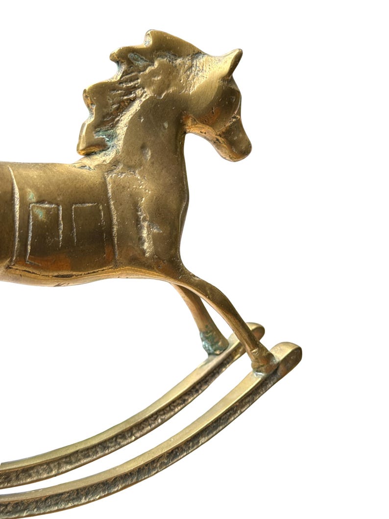 Puede incluir: Una figura de caballo balanc&iacute;n de lat&oacute;n con un acabado dorado met&aacute;lico que presenta p&aacute;tina y oxidaci&oacute;n visibles. El caballo est&aacute; representado en una pose de galope con crines texturizadas, un detalle de silla de montar y dos balancines.