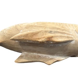 May include: A light brown, hand-carved wooden fish sculpture featuring a smooth, natural grain texture. The fish is depicted in a horizontal swimming position with visible carved details on the fins, tail, and body, showcasing a raw finish.