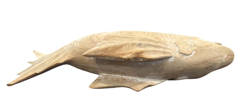 May include: A light brown, hand-carved wooden fish sculpture featuring a smooth, natural grain texture. The fish is depicted in a horizontal swimming position with visible carved details on the fins, tail, and body, showcasing a raw finish.