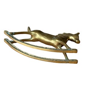 Puede incluir: Una figura de lat&oacute;n pulido con forma de caballo balanc&iacute;n. El caballo aparece en una pose de galope, unido a dos balancines curvos. La superficie met&aacute;lica muestra un tono dorado c&aacute;lido con sutiles variaciones en la textura y el reflejo.