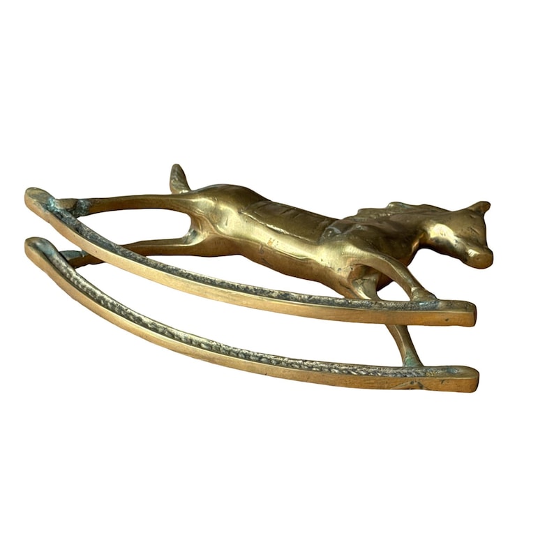 Puede incluir: Una figura de lat&oacute;n pulido con forma de caballo balanc&iacute;n. El caballo aparece en una pose de galope, unido a dos balancines curvos. La superficie met&aacute;lica muestra un tono dorado c&aacute;lido con sutiles variaciones en la textura y el reflejo.