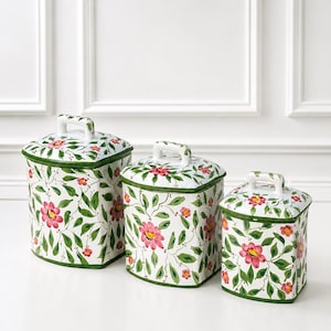 May include: A set of three white ceramic canisters with lids, each featuring a hand-painted floral pattern with pink flowers, green leaves, and thin brown vines. The canisters have a square shape with rounded corners and green trim.