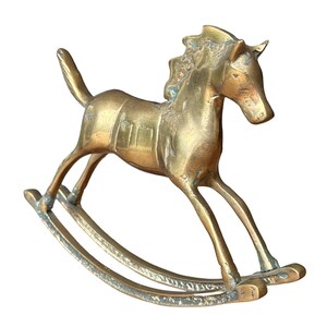 Puede incluir: Una figura de caballo balanc&iacute;n de lat&oacute;n con acabado dorado met&aacute;lico. El caballo presenta una crin texturizada, un detalle de silla de montar en el lomo y una cola levantada. Est&aacute; montado sobre dos balancines curvos y muestra una p&aacute;tina envejecida.