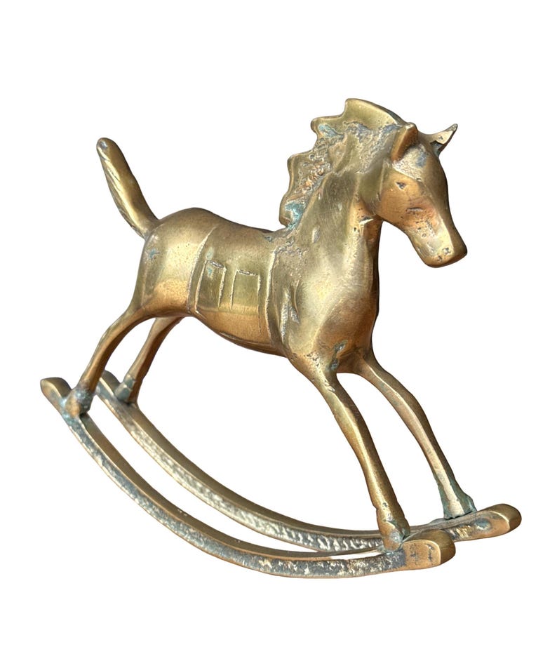 Puede incluir: Una figura de caballo balanc&iacute;n de lat&oacute;n con acabado dorado met&aacute;lico. El caballo presenta una crin texturizada, un detalle de silla de montar en el lomo y una cola levantada. Est&aacute; montado sobre dos balancines curvos y muestra una p&aacute;tina envejecida.