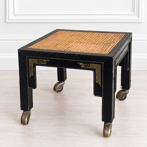 May include: A square black wooden side table featuring a natural woven cane top insert. The frame is accented with gold-toned corner detailing and is supported by four straight legs, each equipped with a functional metal caster wheel for mobility.