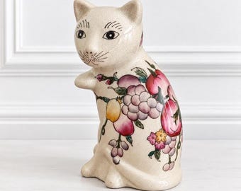 Peut inclure: Une figurine de chat en céramique de couleur crème avec une finition émaillée craquelée. Le chat est orné d'un motif peint coloré représentant divers fruits, notamment des raisins, des pêches et des baies, ainsi que des feuilles vertes.