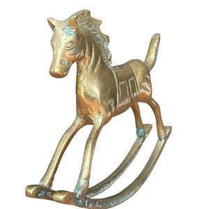 Puede incluir: Una figura de caballo balanc&iacute;n de lat&oacute;n con un acabado dorado met&aacute;lico y p&aacute;tina visible. El caballo presenta una crin esculpida, un detalle de silla de montar en el lomo y una cola hacia arriba, montado sobre dos balancines curvos.
