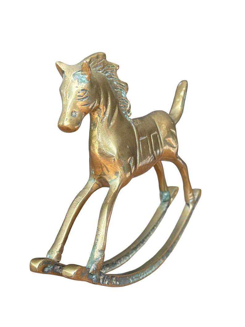Puede incluir: Una figura de caballo balanc&iacute;n de lat&oacute;n con un acabado dorado met&aacute;lico y p&aacute;tina visible. El caballo presenta una crin esculpida, un detalle de silla de montar en el lomo y una cola hacia arriba, montado sobre dos balancines curvos.