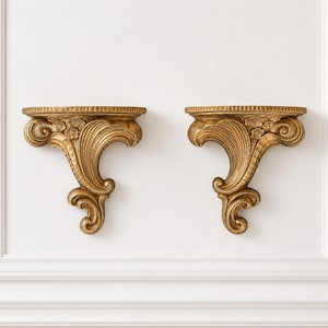 May include: A pair of gold-colored wall shelves featuring ornate, carved scrollwork and floral detailing. Each shelf has a flat, semi-circular top surface supported by a decorative bracket with curved, sweeping lines and intricate relief patterns.