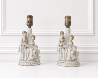 Peut inclure: Une paire de lampes de table en porcelaine représentant un couple sculpté en tenue du XVIIIe siècle. Les figurines sont finies en blanc avec des accents dorés et des détails floraux. Chaque base de lampe supporte une douille en laiton.
