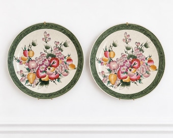 Peut inclure: Une paire d'assiettes rondes en céramique présentant une illustration centrale de fruits et fleurs assortis dans des tons de rouge, rose, jaune et vert, entourée d'une bordure décorative vert foncé avec un motif de feuilles et des attaches en métal doré.