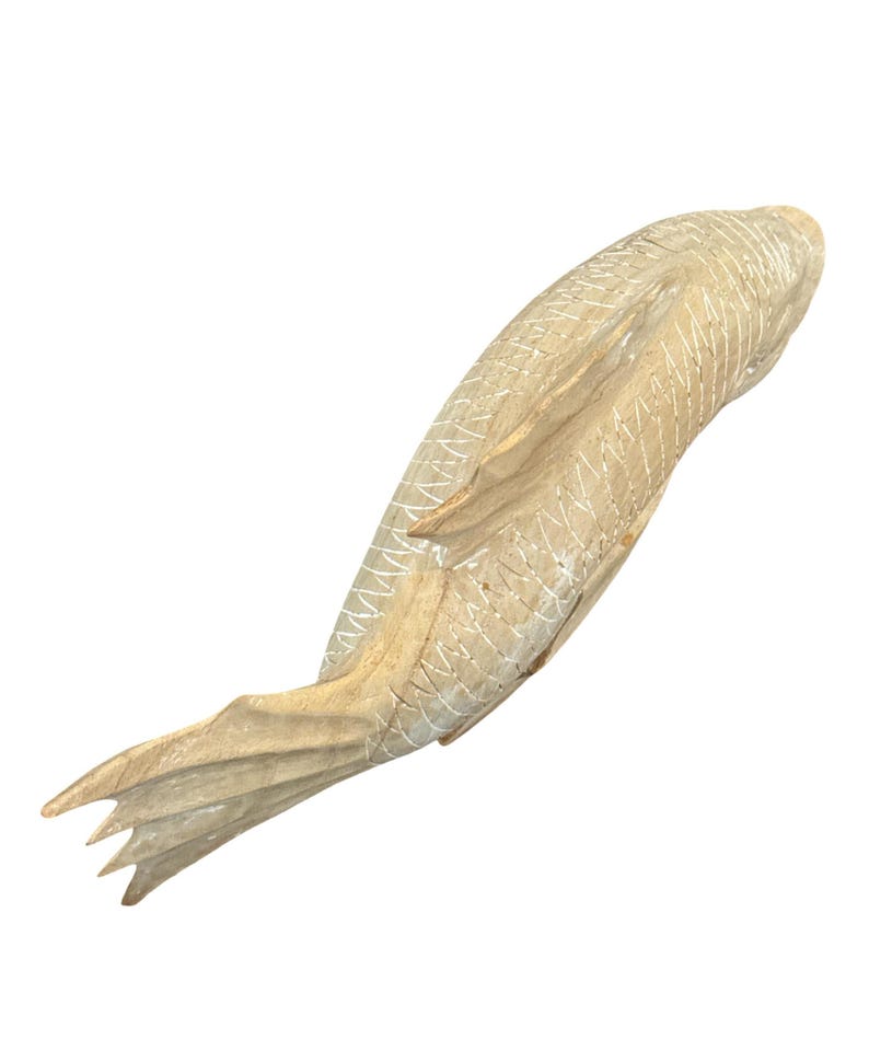 May include: A light brown wooden fish carving featuring a smooth, polished finish and detailed, incised cross-hatch patterns along the body to represent scales. The sculpture displays a curved, elongated shape with a defined tail and fins.