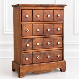 May include: A tall wooden apothecary chest featuring sixteen individual drawers arranged in four rows. Each drawer is equipped with a small, round, white ceramic knob with a black center dot. The cabinet has a warm brown finish and a scalloped base.