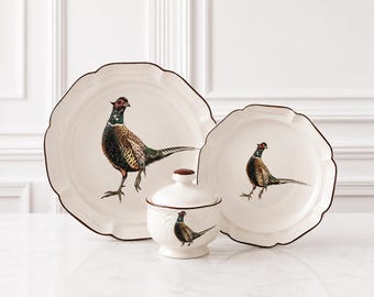 May include: Two cream-colored ceramic plates with scalloped edges and brown rims, each featuring a detailed illustration of a pheasant. A matching lidded sugar bowl with a brown knob and a pheasant illustration sits in the foreground.