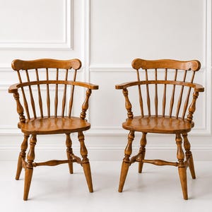 May include: A pair of matching wooden armchairs featuring a medium brown finish, curved backrests with vertical spindles, rounded armrests, and turned legs connected by horizontal stretchers. The chairs have contoured seats and a sturdy frame.