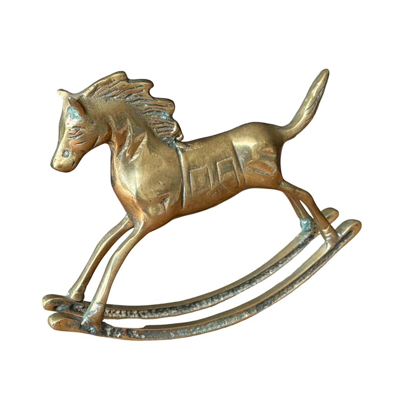 Puede incluir: Una figura de caballo balanc&iacute;n de lat&oacute;n pulido con una crin detallada, un dise&ntilde;o de silla de montar en el lomo y una cola levantada. El objeto met&aacute;lico tiene un tono dorado c&aacute;lido con sutiles signos de p&aacute;tina envejecida en los balancines.