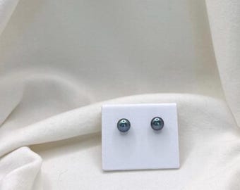 Puede incluir: Un par de pendientes de perlas grises oscuras exhibidos en una tarjeta blanca. Los pendientes son redondos y tienen un brillo sutil. El fondo es una tela blanca texturizada.