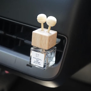 Op de afbeelding: Een auto-geurverfrisser met een heldere glazen fles en een houten bovenkant. De houten bovenkant heeft twee kleine, ronde, lichtgekleurde houten accenten. Het label leest "EUCALYPTUS + MINT" en "car diffuser".