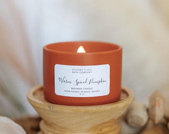 May include: A lit, orange-colored candle in a ceramic container, labeled "Warm Spiced Pumpkin." The candle sits on a wooden stand. The text "Hickory Hill Bath Company" is also visible. The candle is 5 oz.