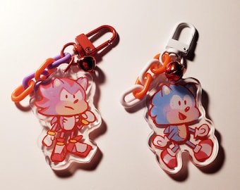 May include: Two acrylic keychains featuring cartoon characters. One keychain has a red clasp, purple and orange links, and a red bell, with a pink character. The other has a white clasp, white and orange links, and a red bell, with a blue character.