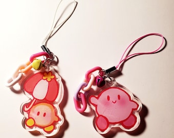 May include: Two acrylic charms featuring the character Kirby. One charm shows Kirby holding a red and white umbrella with a white cord. The other charm shows Kirby with a pink cord. Both include small plastic chain links and tiny metal bells.