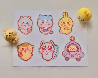 May include: A sheet of colorful cartoon stickers featuring various cute animal characters, including a bear, cat, rabbit, and squirrel. The stickers have red outlines and are in a kawaii style. Two yellow figurines of a rabbit and a round character are also present.