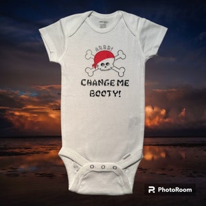 May include: White baby onesie with a pirate-themed graphic. The design features a skull wearing a red bandana, crossed bones, and the text "ARRR!" and "CHANGE ME BOOTY!" in black lettering. The onesie has short sleeves and snap closures.