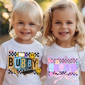 May include: Two children wearing white t-shirts with graphic designs. The shirt on the left says "Bubby" in colorful block letters with a checkered background and a skateboard graphic. The shirt on the right says "Sissy" in colorful block letters with a checkered background and a pink bow.