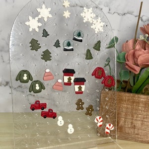 May include: A clear acrylic display stand adorned with festive Christmas-themed charms. The charms include snowflakes, Christmas trees, snow globes, sweaters, hats, coffee cups, gingerbread men, red trucks, snowmen, and candy canes.