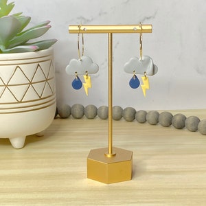 May include: A pair of whimsical earrings featuring gray cloud shapes with blue raindrops and yellow lightning bolts. The earrings hang from gold-colored hoops. The earrings are displayed on a gold-colored stand.
