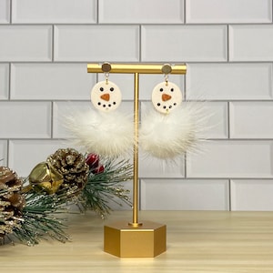 May include: A pair of snowman-themed earrings. Each earring features a cream-colored circular face with black dot eyes and a brown triangle nose. Below the face is a fluffy white feather ball. The earrings hang from a gold-colored post.