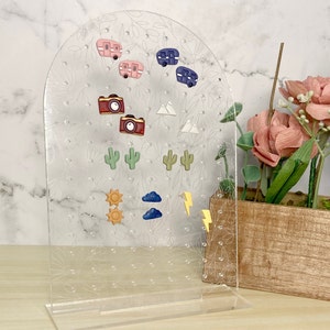 May include: Clear acrylic earring display stand with a floral design. The stand holds various stud earrings, including pink and blue camper trailers, cameras, cacti, suns, clouds, and lightning bolts.