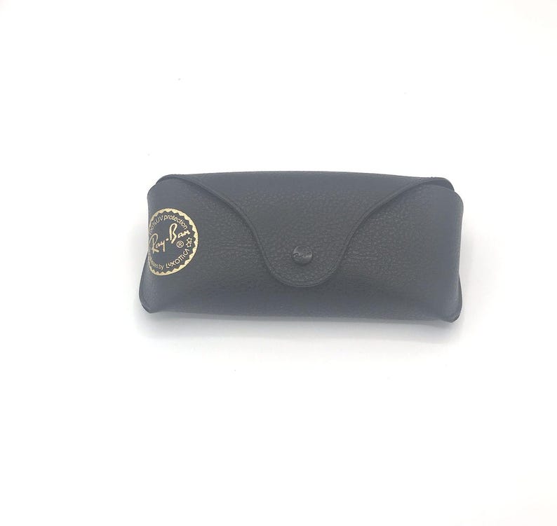 May include: A black, textured sunglasses case with a snap closure. The case features a gold emblem with the words "Ray-Ban" in a circular design. The case is designed to protect eyewear.