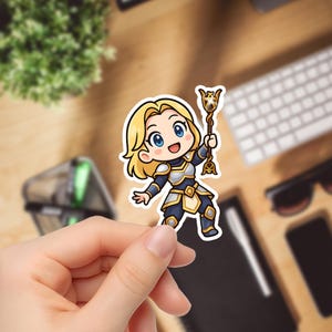 May include: A sticker depicting a cartoon character with blonde hair, blue eyes, and a cheerful expression. The character wears blue and gold armour and holds a golden staff. The sticker is held by a hand against a blurred background.