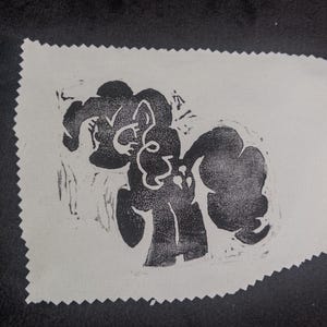 May include: A rectangular piece of off-white fabric with pinked edges featuring a black ink block print of a stylized pony character with a curly mane and tail. The design includes three small heart-shaped symbols on the pony's flank.