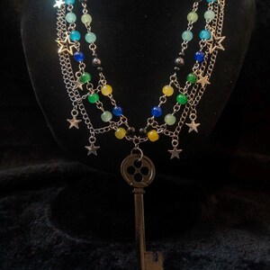 May include: A multi-strand necklace with silver chains, colorful beads, and star charms. The necklace features a large key pendant. The beads are in shades of blue, green, and yellow. The necklace is displayed against a black background.