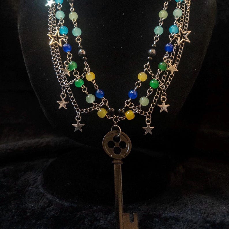 May include: A multi-strand necklace with silver chains, colorful beads, and star charms. The necklace features a large key pendant. The beads are in shades of blue, green, and yellow. The necklace is displayed against a black background.
