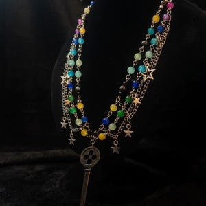 May include: A multi-strand necklace with colorful beads and silver-tone chains. The necklace features a large key pendant and small star charms. The beads are in shades of blue, green, yellow, pink, and black.