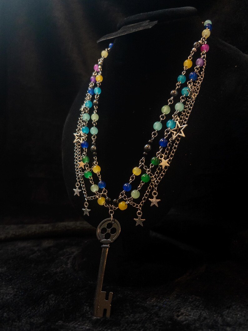 May include: A multi-strand necklace with colorful beads and silver-tone chains. The necklace features a large key pendant and small star charms. The beads are in shades of blue, green, yellow, pink, and black.