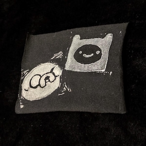 May include: A rectangular black fabric patch featuring a white screen-printed graphic of the characters Finn and Jake from Adventure Time. The design shows the square-headed human character and the round dog character in a stylized, textured ink.