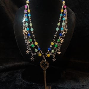 May include: A multi-strand necklace with colorful beads and silver chains. The necklace features a large key pendant and star charms. The beads are in shades of blue, green, yellow, and pink. The necklace is displayed on a black mannequin.