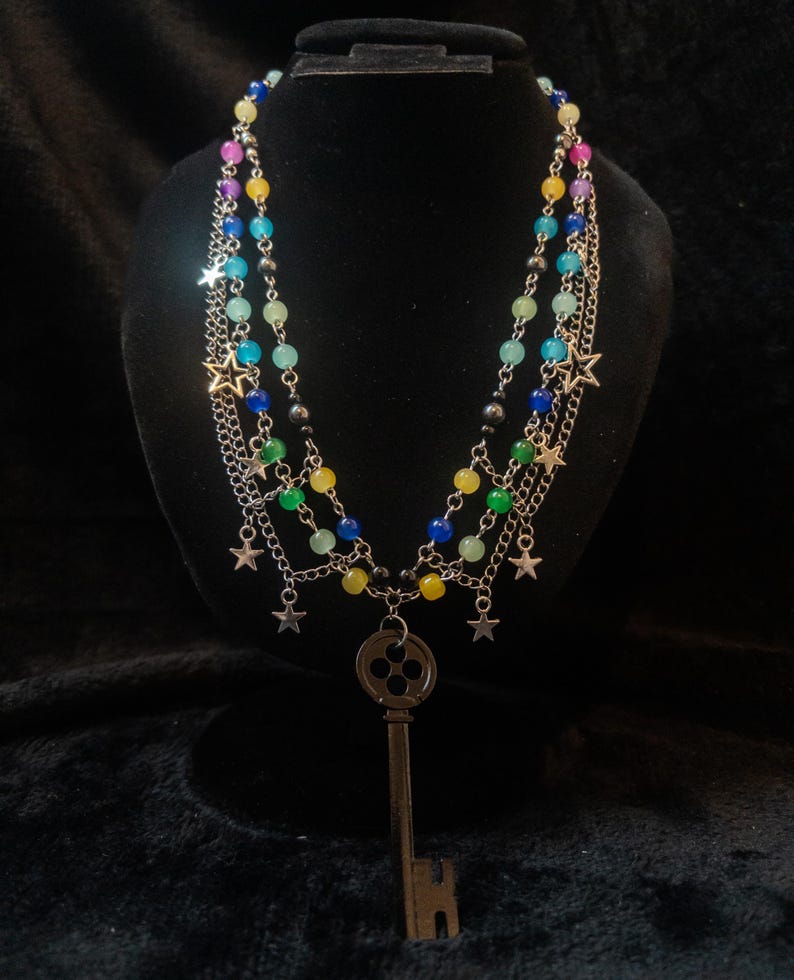 May include: A multi-strand necklace with colorful beads and silver chains. The necklace features a large key pendant and star charms. The beads are in shades of blue, green, yellow, and pink. The necklace is displayed on a black mannequin.