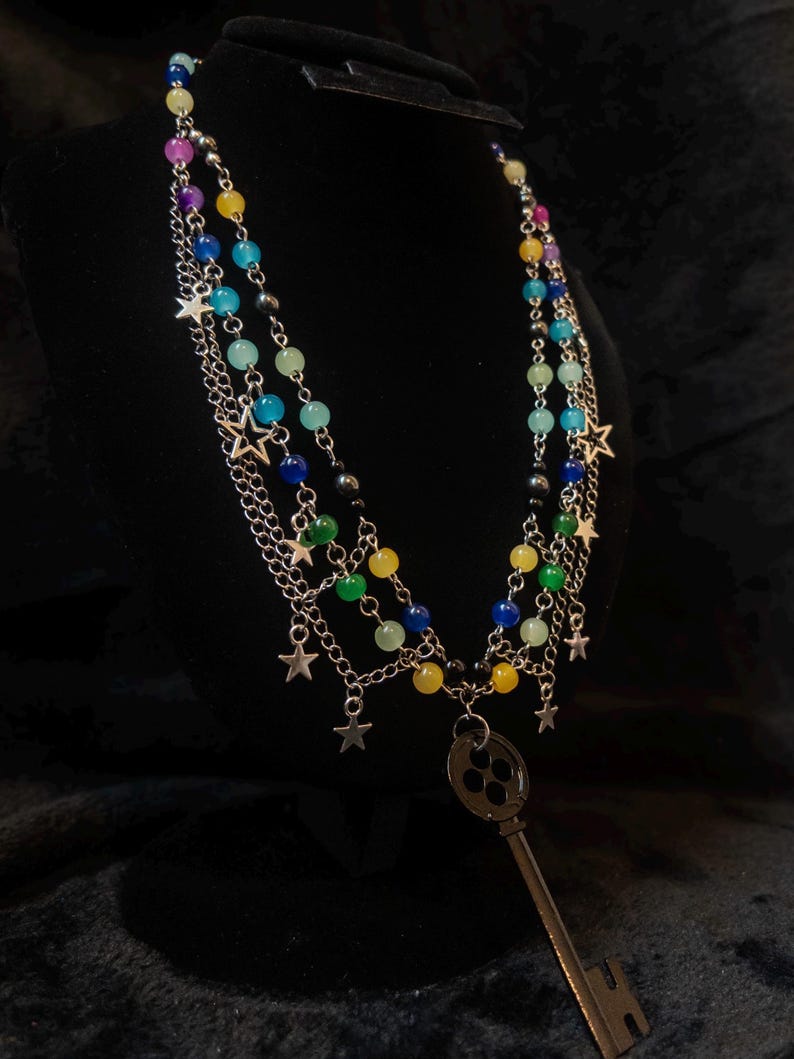 May include: A multi-strand necklace with colorful beads in shades of blue, green, yellow, purple, and black. Silver chains and star charms dangle from the strands. A large, antique-style key pendant hangs from the center.