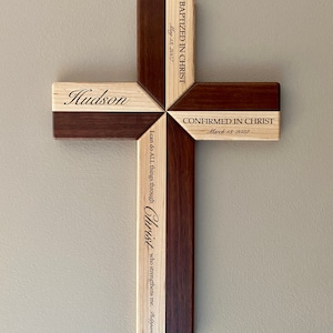 Op de afbeelding: Een houten kruis met een lichtbruin en donkerbruin houten ontwerp. Het kruis is gegraveerd met de naam "Hudson", de data "13 mei 2007" en "13 maart 2022", en de tekst "BAPTIZED IN CHRIST", "CONFIRMED IN CHRIST", en "Ik kan alles door Christus, die mij kracht geeft. Filippenzen 4:13".
