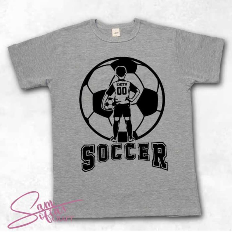 May include: Gray t-shirt with a black soccer ball graphic. The graphic features a young soccer player standing in front of the ball with the text "SOCCER" below. The player's jersey has the text "SMITH" and the number "00" on it.