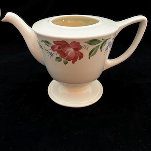 May include: White teapot with a floral design featuring pink roses, green leaves, and blue flowers. The teapot has a footed base.