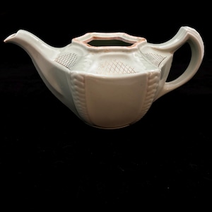 Può includere: Una teiera in ceramica verde pallido con un design geometrico. La teiera ha un beccuccio, un manico e un motivo decorativo sul corpo. Il bordo ha un colore marrone a contrasto.