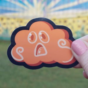 May include: An orange cloud sticker with a surprised expression, outlined in black. The cloud has large, wide eyes and a surprised open mouth. White swirls accent the cloud's design. The sticker is held up against a blurred background.