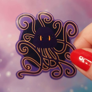 May include: Purple and gold sticker with a black, cartoon-like creature with orange eyes and many tentacles. The creature is surrounded by a swirling, gold pattern.